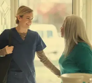 Caregiver in scrubs greeting an older woman at the door with a warm smile.