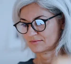 Woman with gray hair and glasses looking down thoughtfully, with a calm expression.