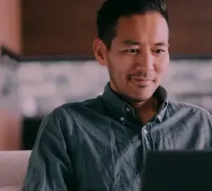 Man sitting on a couch smiling while working on a laptop.