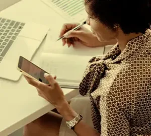 Person writing notes at a desk with a laptop and smartphone.