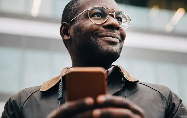 Man outdoors holding a smartphone and looking off to the side with a slight smile.