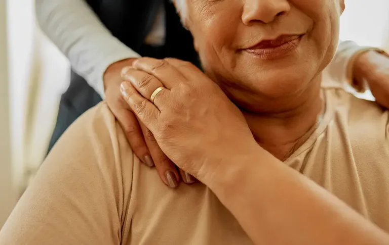 Close-up of an older woman’s hands resting on her shoulder with another person’s hand offering support.