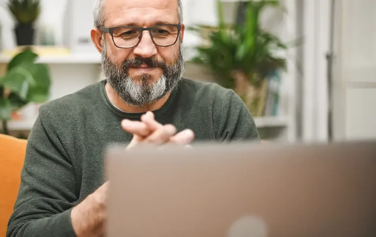 Man with glasses and a beard smiling while sitting in front of a laptop.