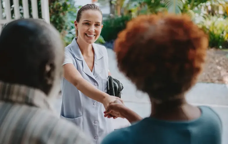 Care worker smiling while shaking hands with an older adult at the doorway.