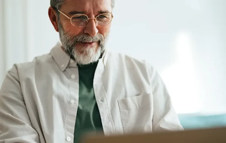 Older man with glasses and a beard smiling while using a laptop.