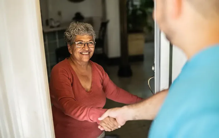 Older woman wearing glasses smiling while shaking hands at her doorway.
