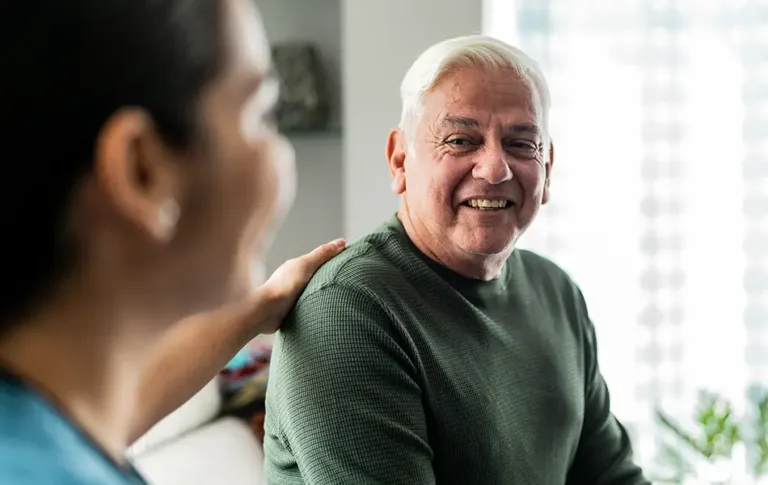 Older man smiling while sitting indoors with a caregiver nearby.