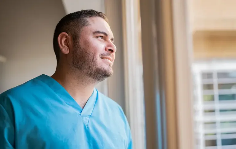 Man wearing blue scrubs smiling while looking out a window.