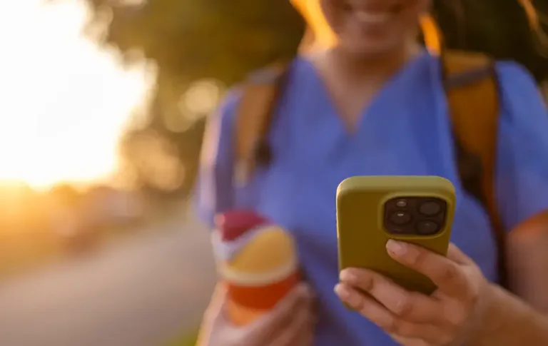 Person in scrubs holding a water bottle and smiling while looking at a smartphone outdoors.