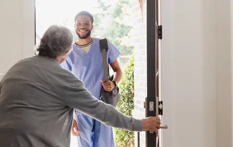 Man in scrubs smiling as he arrives at the door with a bag over his shoulder.