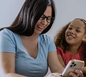 Woman wearing glasses smiling while sitting with a young girl indoors.